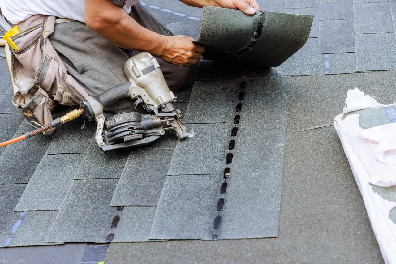 A roofer uses a pneumatic nail gun to install grey asphalt shingles on a roof.