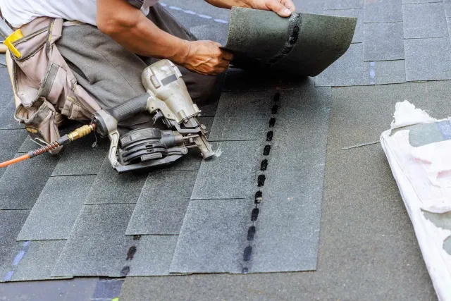 A roofer uses a pneumatic nail gun to install grey asphalt shingles on a roof.