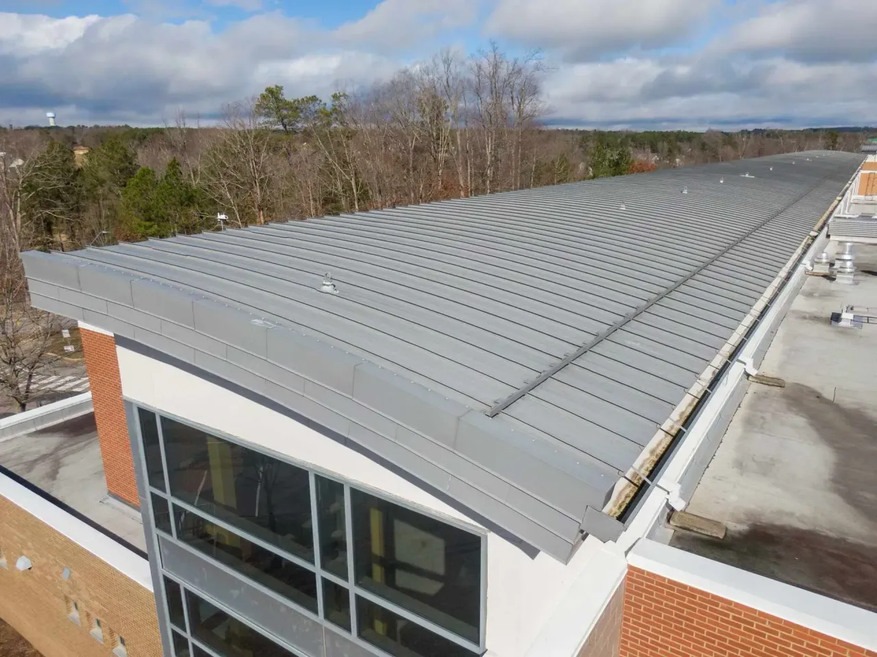 A high-angle view shows a modern building roof featuring a curved, grey standing-seam metal surface against a cloudy sky.