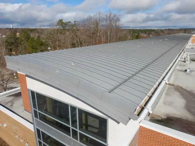 A high-angle view shows a modern building roof featuring a curved, grey standing-seam metal surface against a cloudy sky.