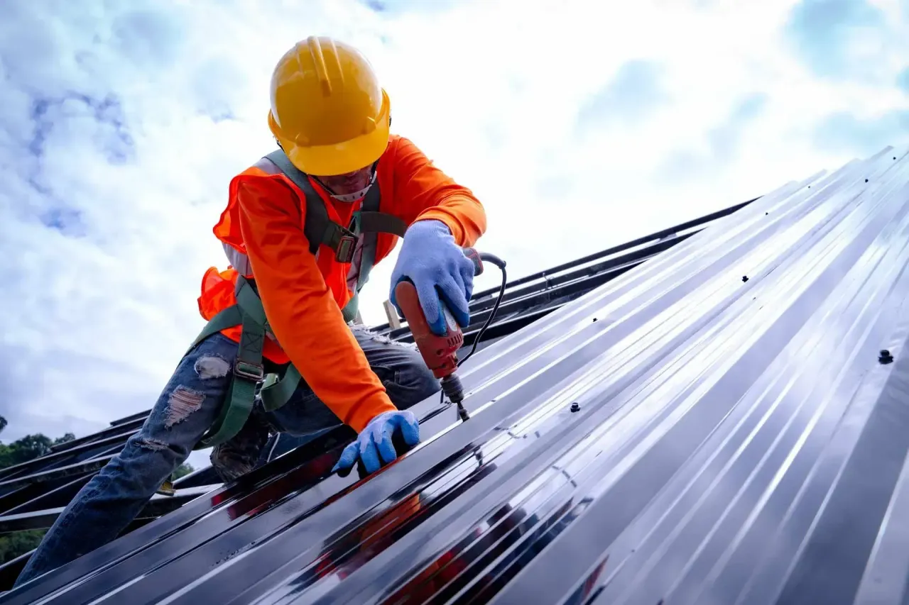 A construction worker in a yellow hard hat and orange shirt uses a drill on a metal roof against a cloudy sky.